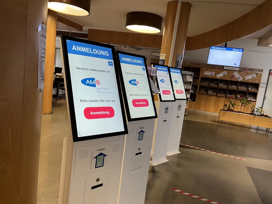 Vier Anmeldeterminals in der Eingangshalle des AMS Wien, jedes mit dem Startbildschirm „Herzlich willkommen — Bitte melden Sie sich an“; im Hintergrund der Wartebereich mit Aufrufmonitor und die Infothek.