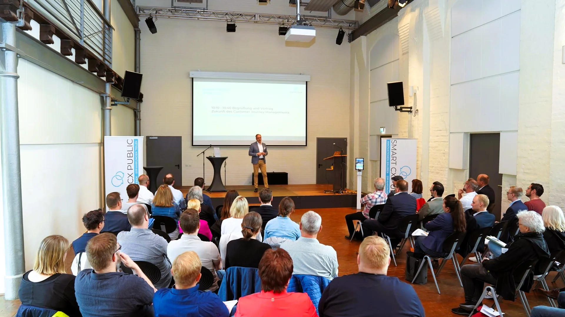 Teilnehmende der Anwendertagung Smart CX Public folgen im Saal einem Vortrag; auf der Bühne spricht ein Referent vor der Projektion des Tagesprogramms.